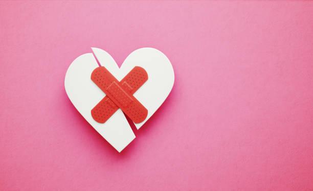 Broken white heart bandaged with red plaster over pink background, Horizontal composition. Broken heart and Valentine's Day concept.